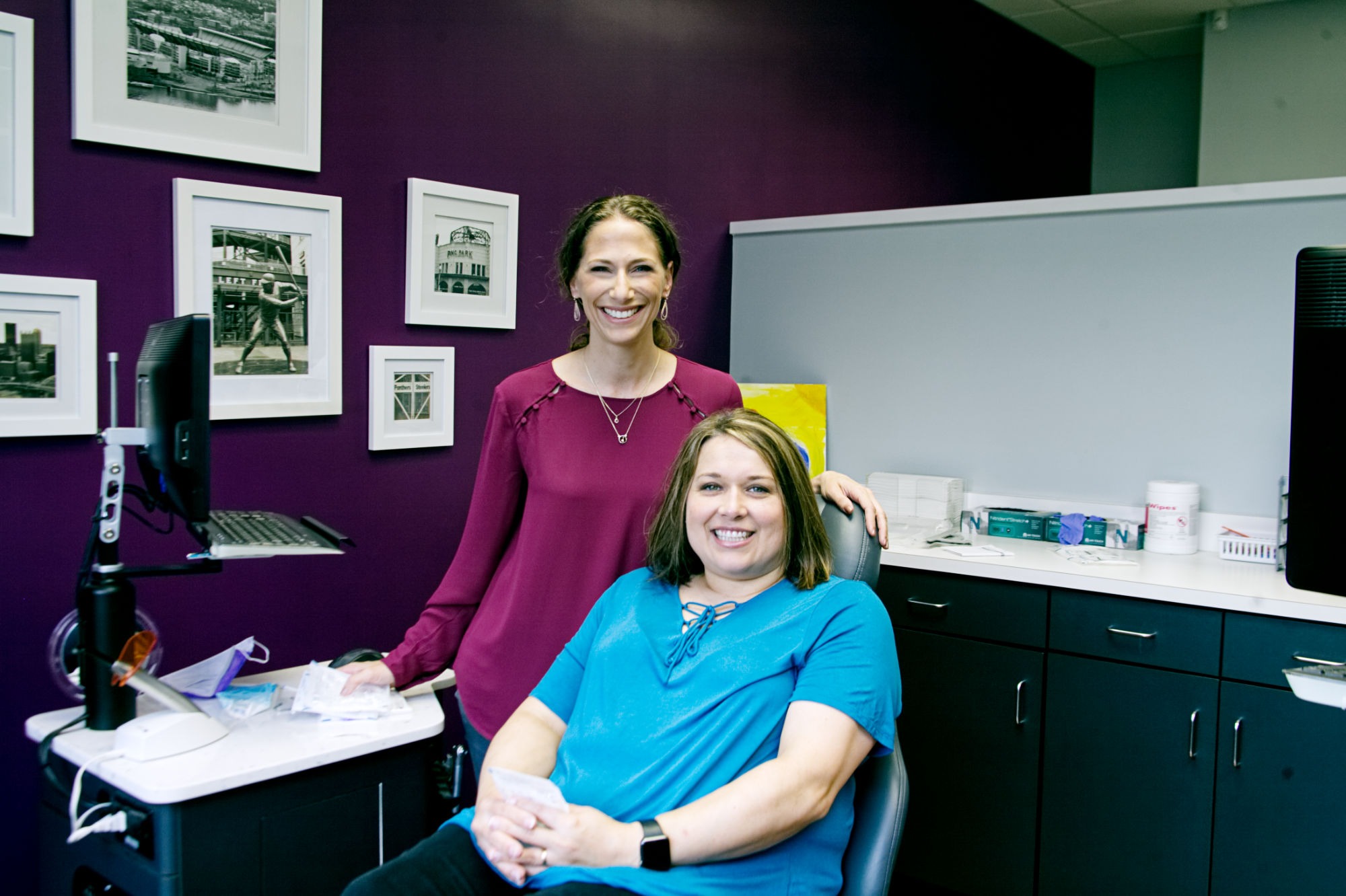 Two smiling team members in a treatment bay at Rhoads Orthodontics, Dr. Stephanie Rhoads’ practice for Mars PA orthodontics, with framed photos on a purple accent wall